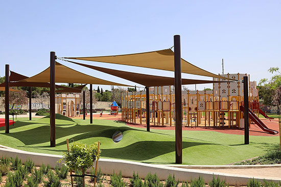 A large playground with a building structure and theme, in the foreground you can see a grass area which is hilly and shaded with tension sail shade installations.